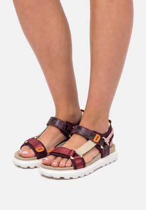 Gambe con sandali bordeaux e beige con cinturini, suole spesse bianche e etichette arancioni con il logo, su sfondo bianco uniforme.