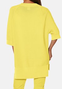 Žlutý oversized pletený top s texturovaným vzorem, krátkými rukávy a bočními rozparky. Design je minimalistický a lehký.