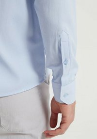 Chemise à boutons bleu clair avec une finition texturée, dotée d'un col pointu et de poignets ajustables, présentée sur le bras et la main d'une personne.