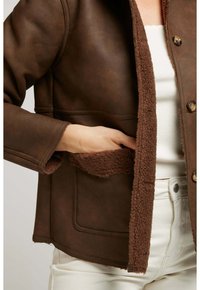 Veste en cuir synthétique marron avec un intérieur en fausse fourrure, fermeture à boutons, poches latérales et une texture lisse, assortie à un pantalon blanc.