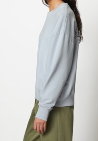 Persona in felpa oversize azzurro chiaro e pantaloni verdi oliva in piedi di profilo con le braccia rilassate su sfondo bianco.