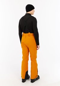 Oranje sneeuwbroek met verstelbare zwarte bretels, met een tapse pasvorm en ritsen bij de onderbenen. Zwarte ski laarzen zijn gedragen.