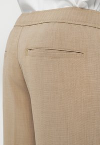 Beige Leinenhosen mit glatter Textur, ausgestattet mit einer Gesäßtasche und einer taillierten Taille. Sichtbare Nähte in einem kontrastierenden Farbton.