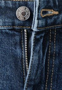 Detailní záběr modrých džínů z denimu, které ukazují kovový knoflík, částečně rozepnutý zip a prošitý knoflíkový otvor béžovou nití.