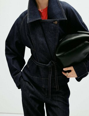 Persona con chaqueta y pantalones vaqueros oscuros, sosteniendo una gran bolsa de cuero negra, mostrando parcialmente una camisa roja debajo.