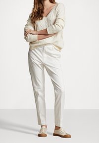 Bílý svetr se strukturovaným pletením, bílé kalhoty na míru a světle béžové tenisky se strukturovanou podrážkou. Uvolněný střih s výstřihem do V a ohrnutými rukávy.