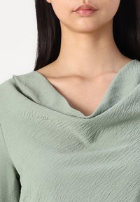 Top verde claro con una tela texturizada y arrugada. Presenta un escote drapeado que se pliega suavemente sobre los hombros.