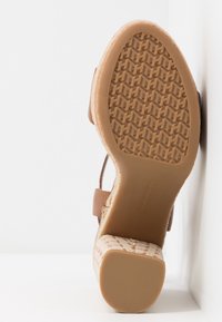 Sandale avec semelle en caoutchouc texturée et talon bloc reposant contre une surface blanche, montrant la tige beige tissée et les détails de la bride cheville.