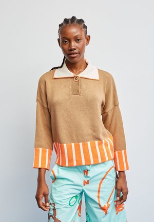 Brauner Strickpullover mit cremefarbenem Kragen und orangefarbener Umrandung, mit einem Knopf und gestreiftem Saum in Orange und Weiß. Kombiniert mit hellblauen Hosen.