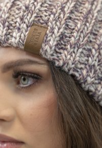Căciulă tricotată în violet deschis și crem, cu un model texturat; prezintă o etichetă din piele maro cu logo. Prim-plan al căciulii și al ochiului.
