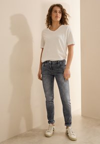 Valkoinen V-pääntie t-paita, haalistuneet harmaat kapeat farkut ja valkoiset tennarit mustilla yksityiskohdilla. Sileä kangas, rento muotoilu, käärityt farkkujen lahkeet.