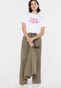 Weißes T-Shirt mit pinkem Grafiktext, kombiniert mit einem hochgeschnittenen, karierter brauner Rock. Das Outfit wird durch weiße Sneaker vervollständigt.