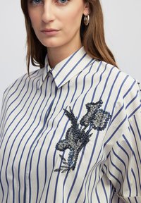Camicia bianca con righe verticali blu, caratterizzata da un design floreale ricamato con paillettes e perline nella zona sinistra del petto.
