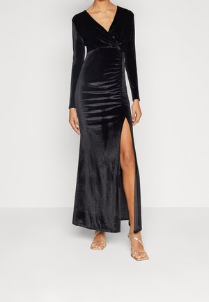 Vrouw in een lange zwarte fluwelen jurk met geplooide details en een hoge split, met gouden sandalen met bandjes, staand tegen een effen achtergrond.