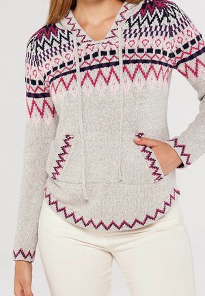 Femme portant un pull en maille gris clair avec des motifs géométriques roses, violets et noirs et un pantalon crème, main dans la poche avant.