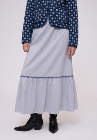 Donna che indossa un giubbotto blu a fantasia, una lunga gonna azzurro chiaro a righe con orlo arricciato e stivali alla caviglia neri a punta.