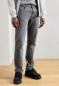 Jeans grigi in denim, caratterizzati da un taglio slim e una texture sottile. Abbinati a scarpe nere in suede con suole testurizzate e lacci.