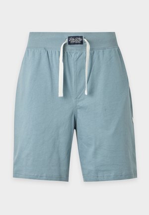 Jeune homme debout pieds nus portant une chemise bleu clair à manches courtes avec de petits motifs blancs et un short assorti avec une poche.