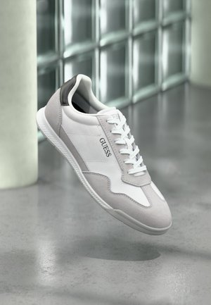 Baskets GUESS blanches et grises avec lacets flottant au-dessus d'un sol gris et d'un mur en blocs de verre translucide en arrière-plan.