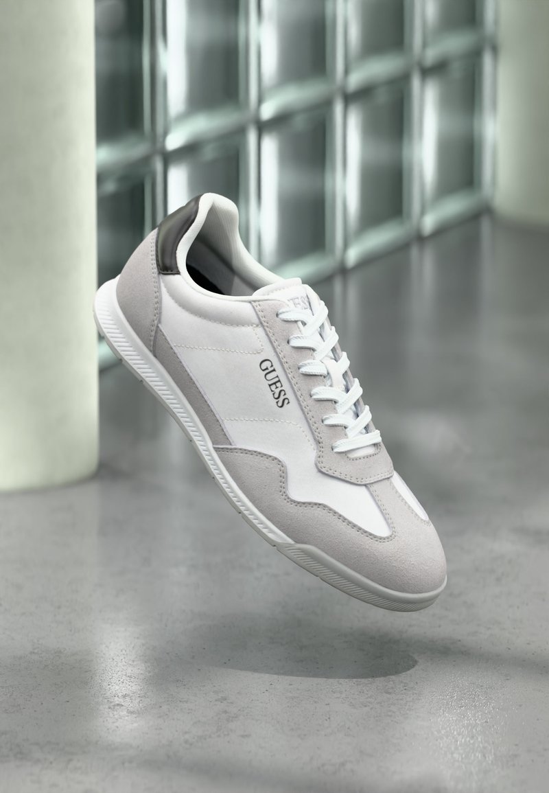 Baskets GUESS blanches et grises avec lacets flottant au-dessus d'un sol gris et d'un mur en blocs de verre translucide en arrière-plan.