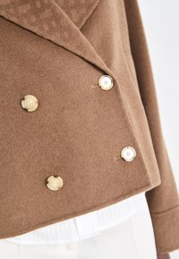 Veste marron à double boutonnage en tissu doux, ornée de boutons en métal doré et d'un col texturé. Superposée sur une chemise blanche plissée.