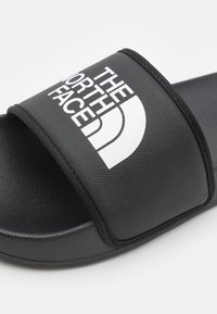 Svart ransle sandal med teksturert stropp og glatt såle. Har hvit logo trykt på stroppen og subtile sømdetaljer.