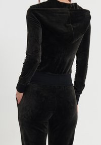 Sudadera con cremallera de terciopelo negro, con capucha, puños y cintura ajustados. Textura suave, diseño elegante, detalles mínimos y sin logotipos visibles.