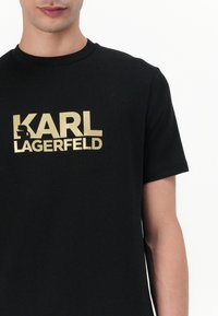 Camiseta de algodón negra con gran texto en oro "KARL LAGERFELD" y un acento gráfico, mangas cortas y cuello redondo.
