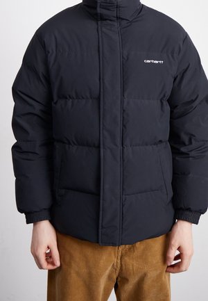 Personne portant une veste matelassée noire Carhartt avec poches avant et un pantalon en velours côtelé marron sur un fond uni.