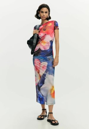 Naine seisab, kandes värvilist lillelist midi-kleiti, musti sandaale ning hoides musta õlakotti lihtsa tausta ees.