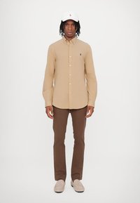 Camisa bege de botão, calças castanhas, boné branco com logótipo e sapatos beges. Tecido suave, ajuste sob medida e design casual.