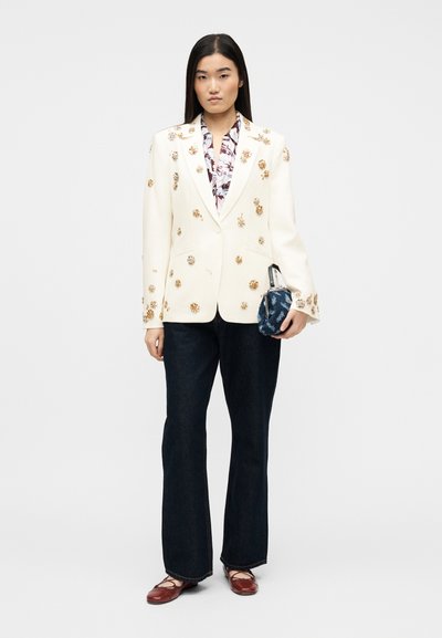 Γυναίκα που φοράει εκρού σακάκι με χρυσά λουλουδένια διακοσμητικά στοιχεία, λουλουδάτο πουκάμισο, σκούρα τζιν με φαρδιά πόδια, κόκκινα παπούτσια και κρατά μια μπλε τσάντα με σχέδια.