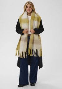 Tørklæde i uldblanding med gule, hvide og brune striber og frynsekant. Modellen bærer en sort oversized jakke og blå vide jeans.