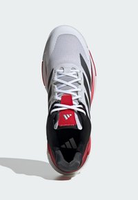 Chaussure de sport blanche avec un dessus en mesh, des accents rouges, un talon noir et trois rayures noires. Elle dispose d'une languette rembourrée et de lacets.
