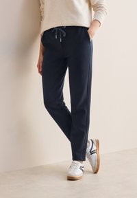 Donkerblauwe joggingbroek van zacht materiaal met een tailleband met trekkoord, zijzakken en taps toelopende pijpen; gecombineerd met witte sneakers.