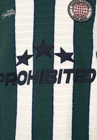 Tela de rayas verticales verdes y blancas con estrellas negras, texto bordado "PROHIBITED", escritura "Berlin Avantgarde" y un parche circular con diseño de globo.
