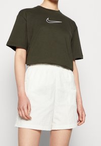 Camiseta de algodón verde oliva con un logotipo blanco de Nike y un corte relajado, combinada con pantalones cortos de lino blancos con cinturilla elástica.
