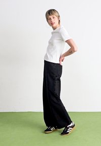 Bijela majica kratkih rukava u kombinaciji s crnim hlačama širokih nogavica i crnim Adidas tenisicama s bijelim prugama. Model stoji bočno na zelenom podu.