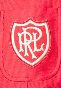 Czerwony materiał z teksturowanym wykończeniem, z białym haftowanym logo tarczy. Logo zawiera splecione litery "R" i "L" w ozdobnym stylu.