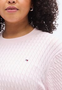 Ροζ ανοιχτόχρωμο πουλόβερ με πλέξη καλωδίου, με στρογγυλή λαιμόκοψη και ριμπ μανσέτες, που διαθέτει ένα μικρό κεντημένο λογότυπο σε κόκκινο, λευκό και μπλε.