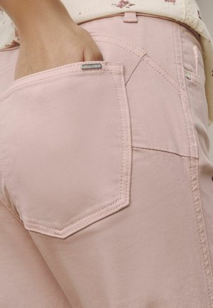 Ruka s prstami vo vnútornom zadnom vrecku svetloružových denimových džínsov, ukazujúca švy vrecka a kovovú značkovú etiketu.