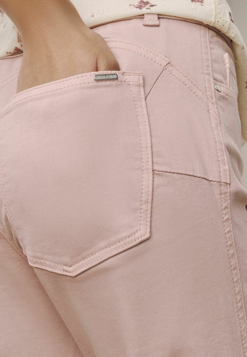 Main avec les doigts dans la poche arrière d'un jean en denim rose clair, montrant les coutures de la poche et une étiquette métallique de la marque.