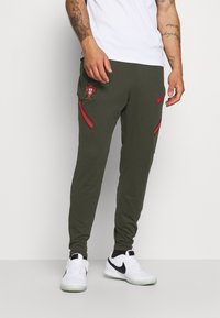 Olivgröna sportbyxor med broderad Portugal-sköld, röda och gröna ränder på sidorna, framfickor och elastiska muddar. Stylat med vita Nike-skor.