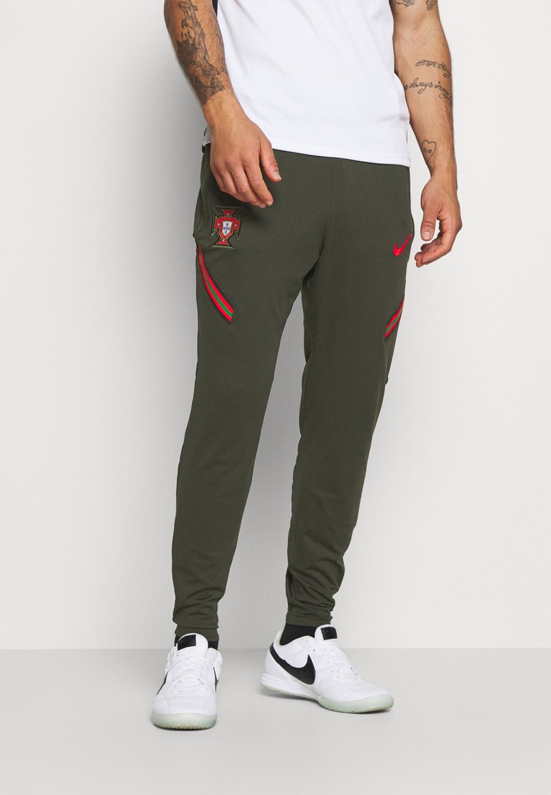 Olivgröna sportbyxor med broderad Portugal-sköld, röda och gröna ränder på sidorna, framfickor och elastiska muddar. Stylat med vita Nike-skor.