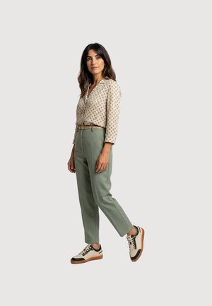Frau steht und trägt eine gemusterte beige Bluse, grüne hoch taillierte Hose mit Gürtel und beige Sneaker mit schwarzen Details vor hellem Hintergrund.
