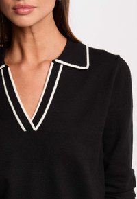 Maglione nero a maglia con scollo a V e colletto bordato di bianco. Il tessuto è liscio con una texture sottile e maniche aderenti.