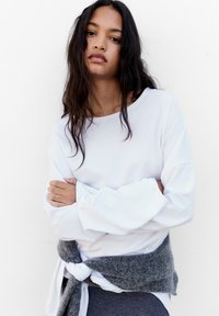 Weißes Langarmoberteil mit weitem Ausschnitt und gerippten Bündchen, kombiniert mit einem geknoteten grauen Pullover, der um die Taille getragen wird. Glatte Stofftextur.