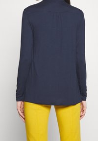 Granatowa bluzka z długim rękawem o luźnym fasonie; miękka tkanina, detal w postaci plis na plecach oraz zaokrąglony dół. Połączona z żółtymi spodniami.