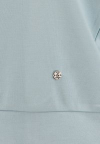 Felpa azzurra chiara realizzata in tessuto morbido, con un distintivo metallico rotondo con un cuore inciso e un testo. Texture semplice e liscia.