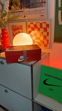 Rød Nike skoeske med hvitt logo, sikret med en brun stropp, hviler på et sølvfarget skap ved siden av en lysende kulelampe og et grønt skilt.